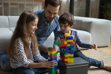 Engaged millennial dad teaching little son and daughter to build tower from construction blocks,...
