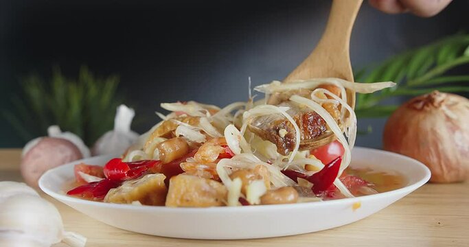 Closeup fresh yummy delicious Thai traditional popular street food spicy green papaya salad or som Tam in white dish, person uses spoon holding crispy pork papaya salad