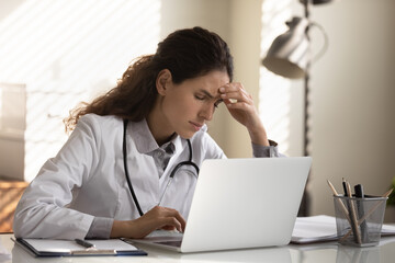 Tired frustrated female doctor feeling stress at workplace, suffering from headache, migraine at computer. Sick exhausted general practitioner holding head at laptop. Medical profession problems