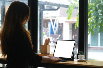 Asian woman typing on tablet and sitting on counter bar beside the window, empty screen of tablet