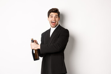 Portrait of attractive man in black suit, winking at camera and opening bottle of champagne,...