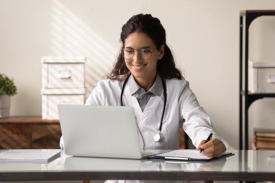 Happy young female GP doctor using laptop, making video call, reading electronic medical records. Medical practitioner giving online virtual consultation to patient from hospital office