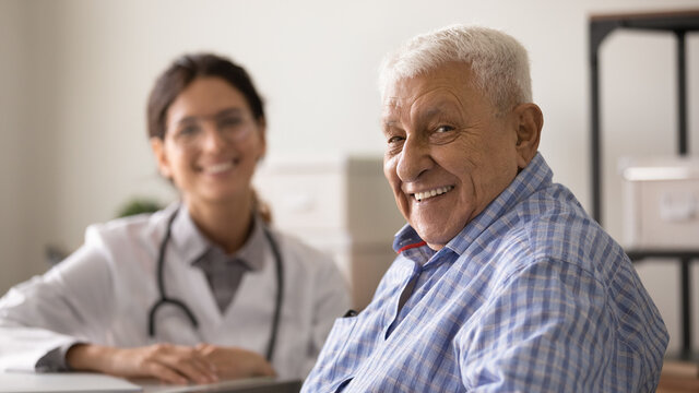 Happy Optimistic Senior 80s Man Looking At Camera With Toothy Smile During Appointment At Doctor Office. Portrait Of Elder Male Patient And Young Female GP Therapist In Background. Medic Care