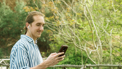 Young handsome hipster man using smartphone and listening music with earphones in the forest