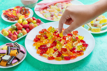 Gummy, jelly, liquorice candies on the table. Hand takes a colorful gummy bear from a plate. An...