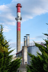 White and red chimney on a background of blue sky and pine trees. Smoking chimney pollution. Gas-powered power plant chimney