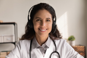 Happy general practitioner in headphones giving online help, medical support, advice, consultation. Head shot portrait of female doctor in headset looking at camera, smiling. video call screen view - obrazy, fototapety, plakaty