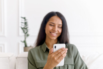 Beautiful young indian woman using smartphone sitting on sofa at home. Charming smiling mixed-race female holds phone and looks at the screen, making video call, watching videos, spends leisure online