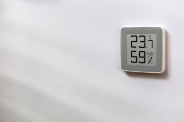 Indoor gray plastic digital thermometer on white wall showing temperature, humidity level, smiling smiley face indoors in daylight sun and parallel stripes of shadow