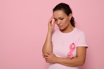 Beautiful thoughtful woman in pink t-shirt with pink ribbon looking down on pink background, hands...