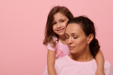 Beautiful baby girl in pink dress with a pink ribbon tenderly hugs her mother with closed eyes, looking at camera, showing support to cancer patients and survivors, pink background with copy space
