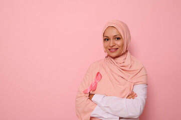 Isolated portrait of Arab Muslim pretty woman wearing pink hijab with pink satin ribbon in symbol of International Day of Cancer Awareness. Oncological diseases concept. Copy space on pink background