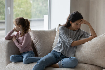 Unhappy young hispanic mother and stressed little child daughter sitting on sofa, ignoring each...