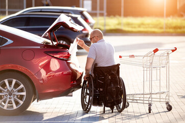 Adult disabled man in a wheelchair puts purchases in the trunk of a car in a supermarket parking lot