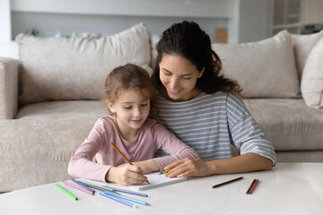 Caring young hispanic woman or babysitter teaching preschool kid girl handwriting letters or drawing in paper copybook, enjoying early development activities, homeschooling domestic study concept.