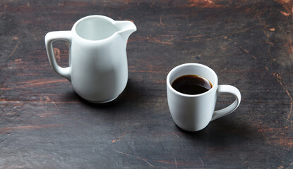 White cup with coffee and milk jug on a white wooden background, closeup. Energy breakfast, morning routine concept