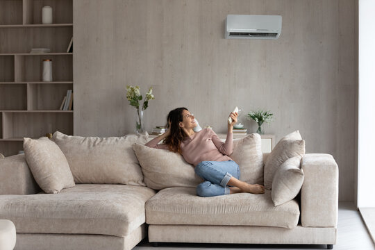 Relaxed Young Hispanic Female Homeowner Sitting On Huge Comfortable Couch, Turning On Air Conditioner With Remote Controller, Switching On Cooler System, Setting Comfortable Temperature In Living Room