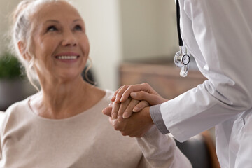 Happy senior woman visiting doctor, getting optimistic news after medical checkup, therapy. Therapist holding hand of old patient, giving hope, support, congratulating on goor treatment result