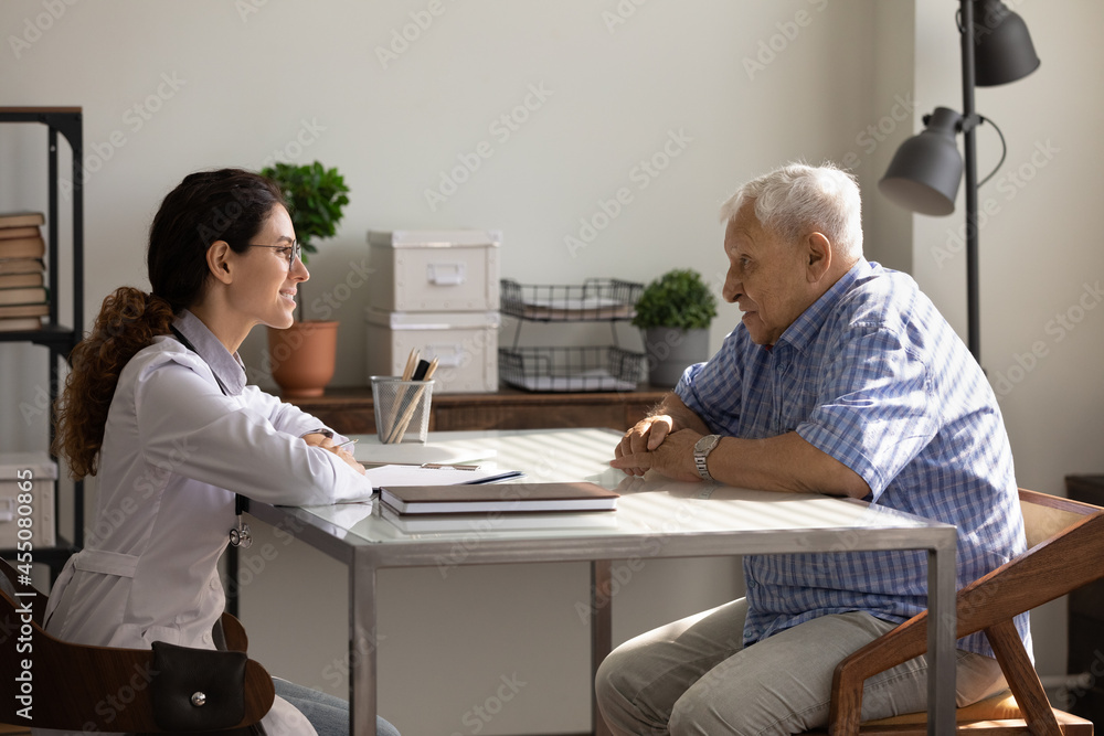 Wall mural Smiling young doctor and mature 80s patient meeting and talking in therapist office. Old man visiting, consulting physician, listening recommendations for treatment and therapy. Geriatric healthcare