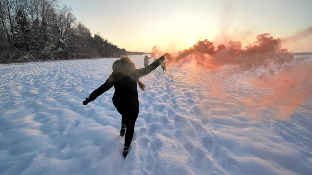 Girls friends run across the winter field with colored smoke.