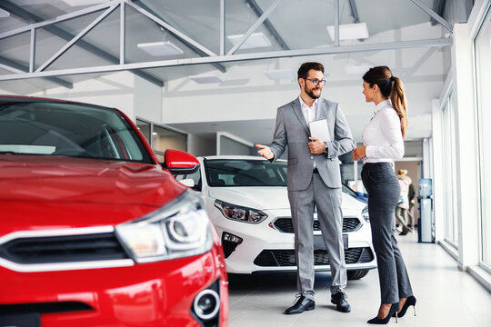 Smiling car seller standing in car salon with customer and showing around cars on sale.