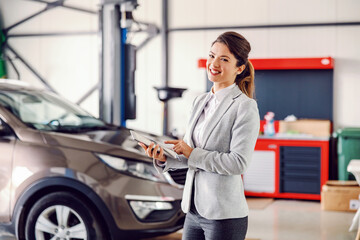 Smiling female car seller standing in garage of car salon and using tablet to check on lager.
