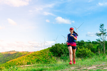 A female trail runner, dressed for runners, sportswear, is practicing on a dirt path in a high mountain forest. with a happy mood, Behind there is a wind turbine that generates electricity.