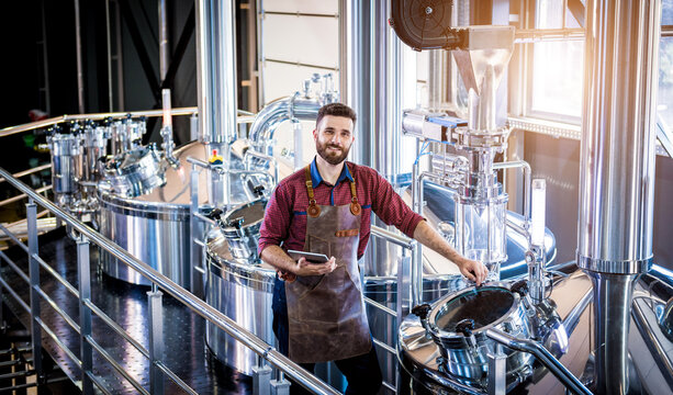 Young male brewer in leather apron supervising the process of beer fermentation at modern brewery factory