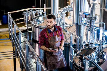 Young male brewer in leather apron supervising the process of beer fermentation at modern brewery...
