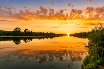 Plakat Scenic view at beautiful summer river sunset with reflection on water with green bushes, calm water ,deep colorful cloudy sky and glow on horizon on a background, spring evening landscape