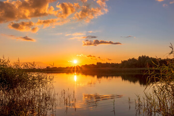 Scenic view at beautiful summer sunset on lake with reflection on water with reeds, grass, golden sun rays, calm water ,deep blue cloudy sky and glow on a background, spring evening landscape