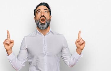 Middle aged man with beard wearing casual white shirt amazed and surprised looking up and pointing with fingers and raised arms.