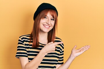 Redhead young woman wearing wool cap amazed and smiling to the camera while presenting with hand and pointing with finger.