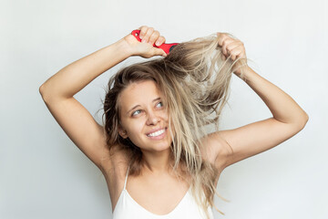 A young blonde woman combing her tangled unruly dry bleached hair with a red comb isolated on a white background. Hair care - obrazy, fototapety, plakaty