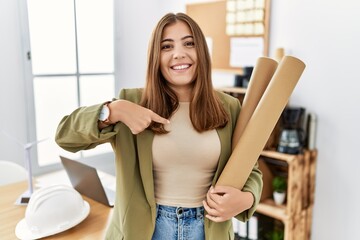 Young brunette woman holding paper blueprints at the office pointing finger to one self smiling happy and proud