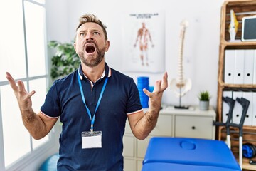 Middle age physiotherapist man working at pain recovery clinic crazy and mad shouting and yelling...