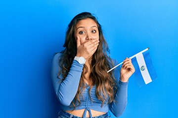 Young hispanic girl holding el salvador flag covering mouth with hand, shocked and afraid for...