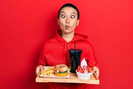Beautiful hispanic woman with short hair eating a tasty classic burger with fries and soda making fish face with mouth and squinting eyes, crazy and comical.