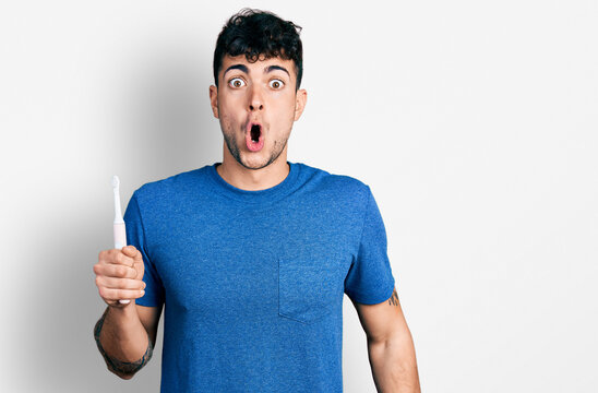 Young hispanic man holding electric toothbrush scared and amazed with open mouth for surprise, disbelief face