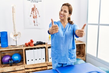 Middle age hispanic physiotherapist woman working at pain recovery clinic approving doing positive gesture with hand, thumbs up smiling and happy for success. winner gesture.