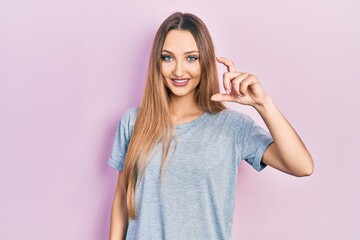 Young blonde girl wearing casual t shirt smiling and confident gesturing with hand doing small size sign with fingers looking and the camera. measure concept.