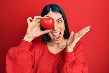 Young hispanic woman holding heart over eye celebrating achievement with happy smile and winner expression with raised hand