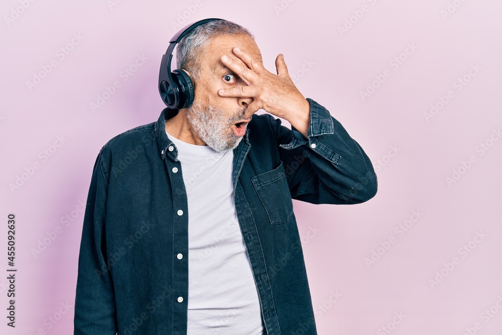 Wall mural handsome senior man with beard listening to music using headphones peeking in shock covering face an