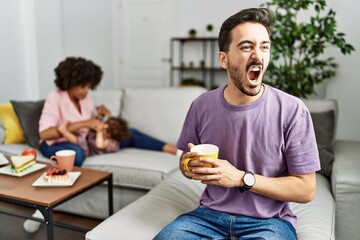 Obraz na płótnie Canvas Hispanic father of interracial family drinking a cup coffee angry and mad screaming frustrated and furious, shouting with anger. rage and aggressive concept.