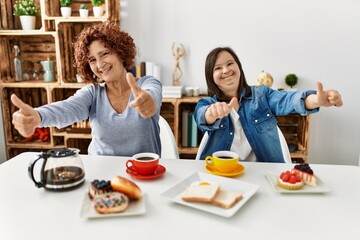 Family of mother and down syndrome daughter sitting at home eating breakfast approving doing positive gesture with hand, thumbs up smiling and happy for success. winner gesture.