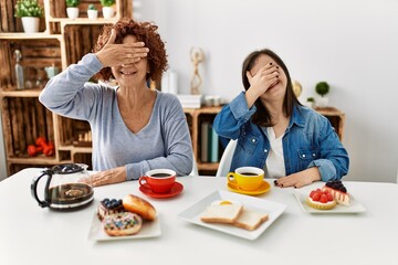 Family of mother and down syndrome daughter sitting at home eating breakfast smiling and laughing with hand on face covering eyes for surprise. blind concept.