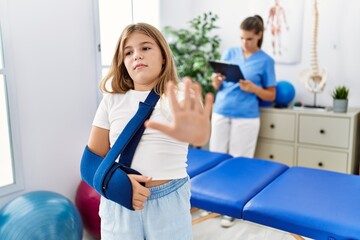 Blonde little girl wearing arm on sling at rehabilitation clinic with open hand doing stop sign with serious and confident expression, defense gesture