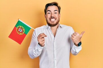 Handsome caucasian man with beard holding portugal flag pointing thumb up to the side smiling happy with open mouth