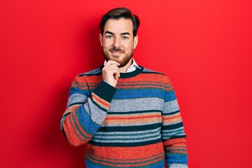 Handsome caucasian man with beard wearing elegant wool winter sweater looking confident at the camera with smile with crossed arms and hand raised on chin. thinking positive.