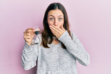 Young brunette woman holding sand clock covering mouth with hand, shocked and afraid for mistake....